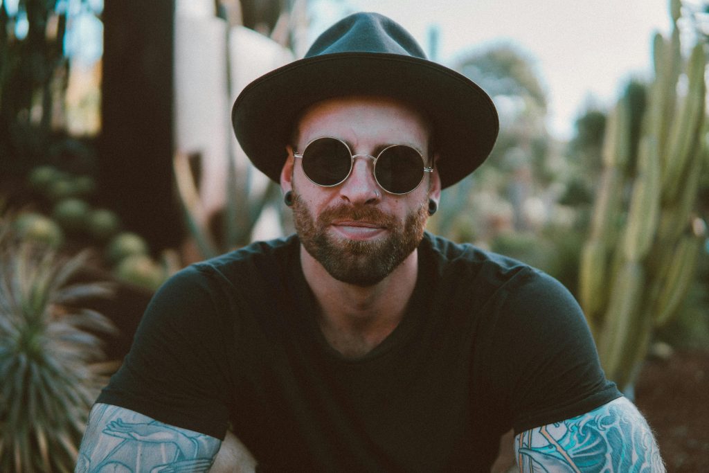 pexels photo 1680172 1680172 Stylish man with beard and tattoos wearing sunglasses and a hat, enjoying outdoors.